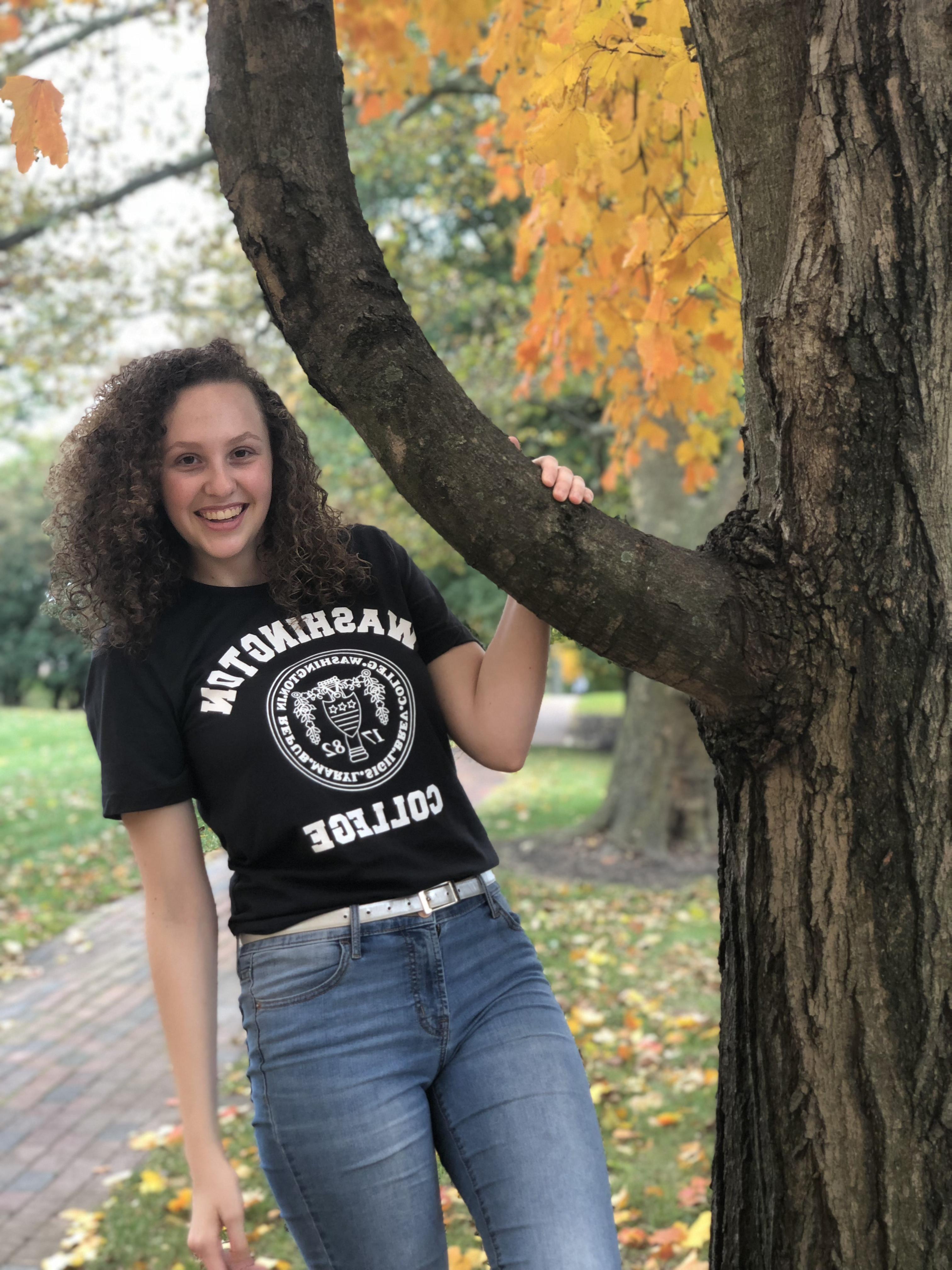 Carlee, smiling 和 wearing a black t-shirt with the 九州娱乐官网 logo, 蓝色牛仔裤, 和 a white belt in front of fall foliage. 